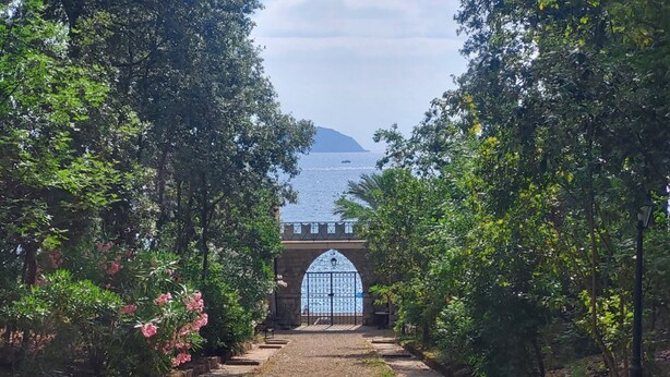 Un sendero conduce a un edificio con una gran entrada arqueada, rodeado de vegetación exuberante y vistas al mar.
