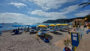 Una playa con filas de tumbonas y sombrillas, un paseo marítimo de madera y vistas al mar y colinas distantes.