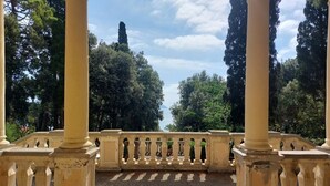 Vista desde un balcón con suelo de baldosas, rodeado de columnas y barandillas, con vista a un parque con árboles y una montaña distante.