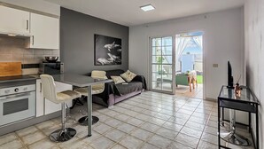 A modern kitchen with white cabinets, a microwave, and a wooden cutting board. There is a dining area with a black sofa and a glass table.