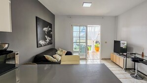 A modern living room with a sofa, a flat-screen TV, a glass coffee table, and a dining area with a black countertop and bar stools.