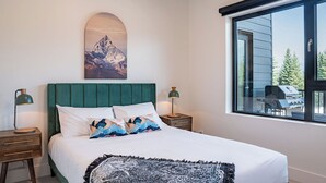 A bedroom with a large bed, a green headboard, two bedside tables with lamps, and a large window with a view of outdoor furniture. - Slopes and Green by Revelstoke Vacations (Revelstoke)