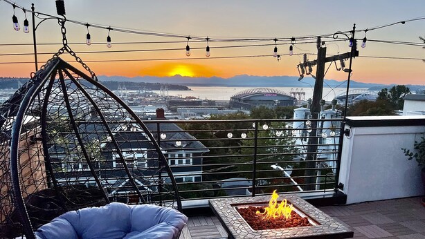 A rooftop terrace with a hanging chair, a fire pit, and a view of a stadium and cityscape during sunset. - Rooftop•Mural•1GB•Parking•AC•FIFAStadiums•W/D•BBQ•Mountain&Bay Views•New Kitchen (Seattle)