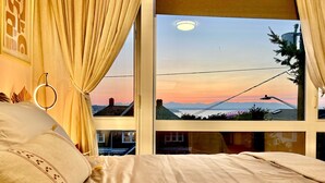 A bedroom with a large window, a bed with white bedding, and a view of a sunset over a cityscape. - Rooftop•Mural•1GB•Parking•AC•FIFAStadiums•W/D•BBQ•Mountain&Bay Views•New Kitchen (Seattle)