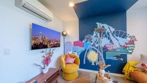 A modern living room with a cityscape painting, a mural of Seattle landmarks, a yellow armchair, and a round coffee table. - Rooftop•Mural•1GB•Parking•AC•FIFAStadiums•W/D•BBQ•Mountain&Bay Views•New Kitchen (Seattle)
