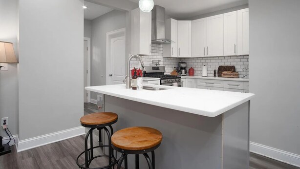 A modern kitchen with white cabinets, a white countertop, two wooden stools, and pendant lighting. - Charming 4 Bedroom Home Near Downtown Columbus with Garage Parking and Backyard. (Columbus)