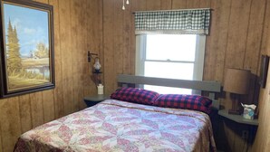 Un dormitorio con cama, mesita de noche, lámpara, cuadro y ventana con cortina a cuadros.
