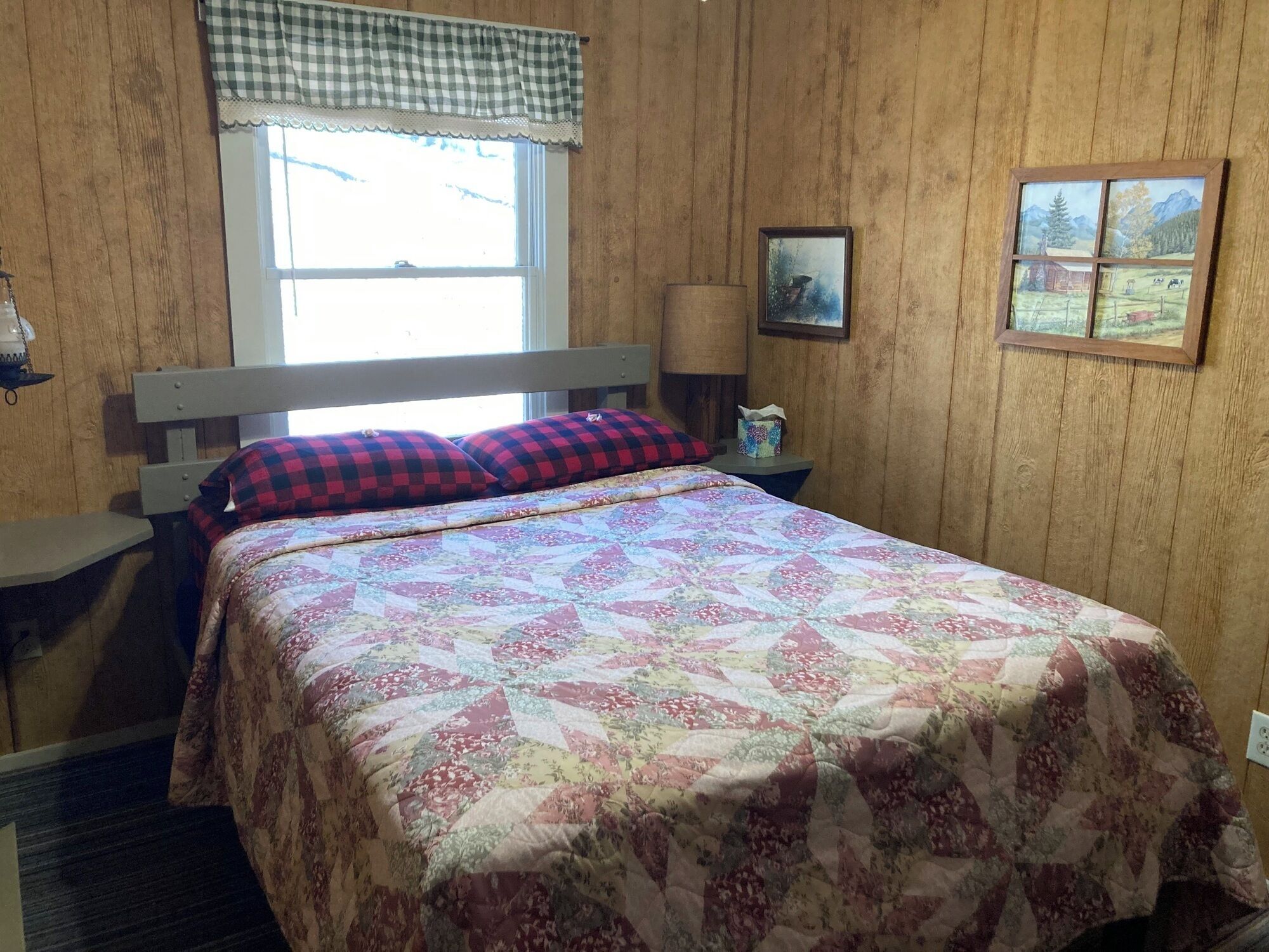 A bedroom with a wooden headboard, a quilted bedspread, a checkered pillow, a bedside lamp, and a framed picture on the wall.