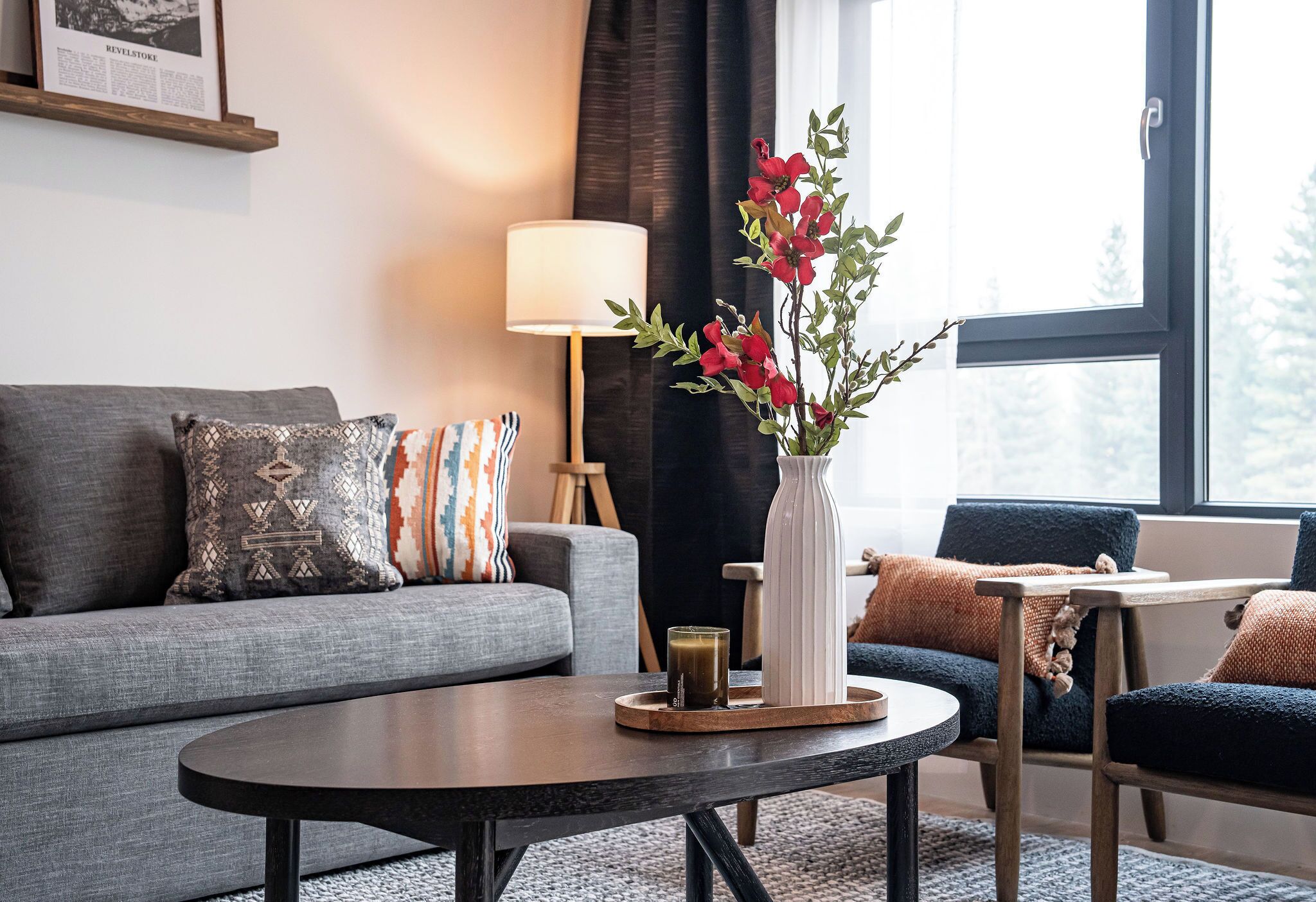 A living room with a grey sofa, patterned pillows, a round coffee table with a vase of red flowers, a lamp, and a window with a view of trees.