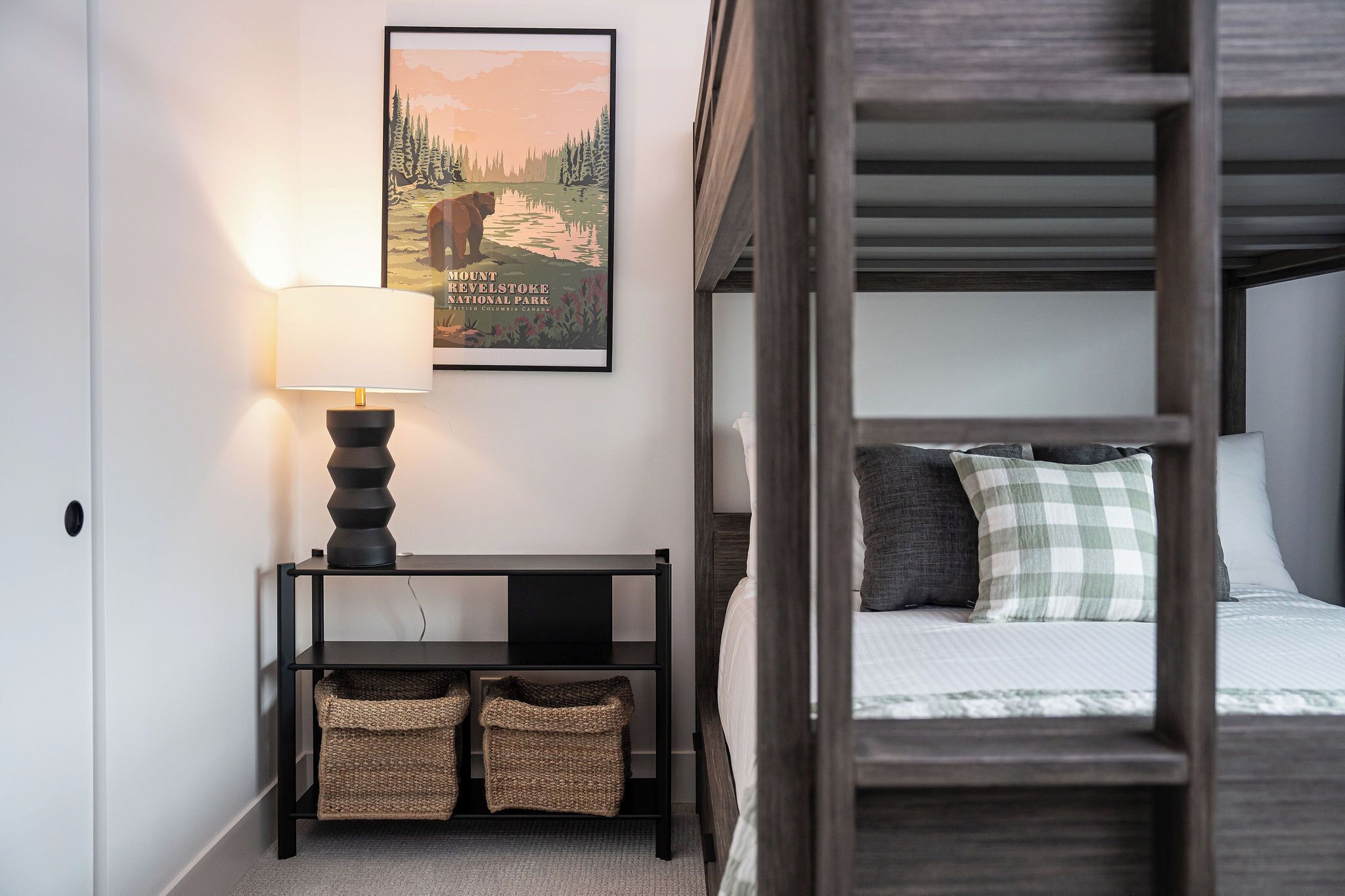 A bedroom with a bunk bed, a bedside table with a lamp, a framed poster on the wall, and two baskets on the floor.