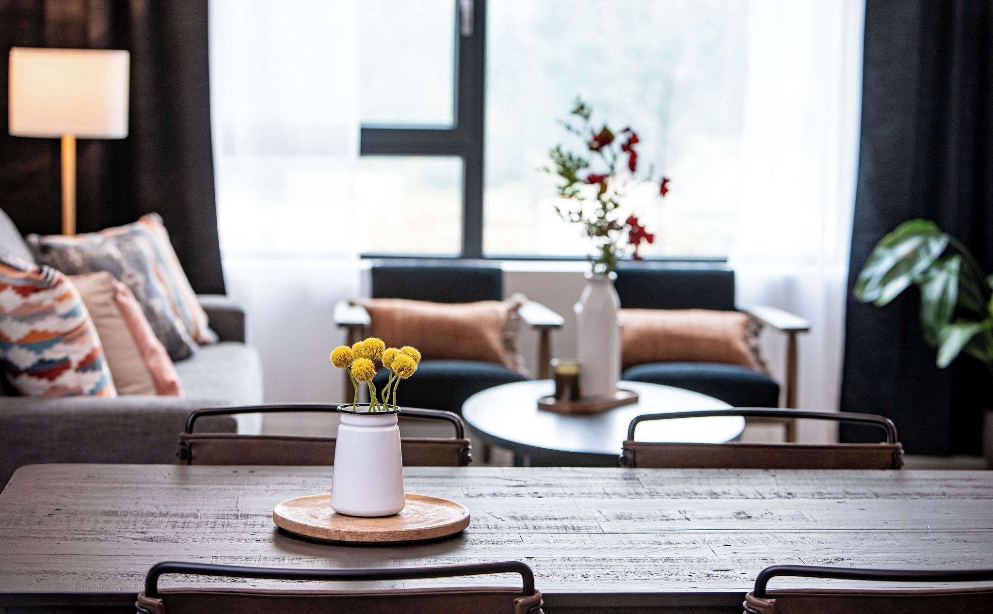 A dining table with a white vase of yellow flowers, a wooden coaster, and a metal chair.