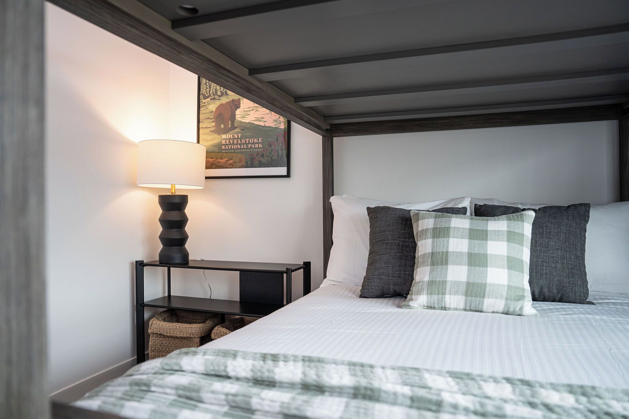 A neatly made bed with a checkered blanket, a bedside lamp, and a framed picture of Mount Revelstoke National Park.