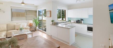 A modern kitchen with white cabinets, a dining area with a wooden table, and a living room with a beige sofa.