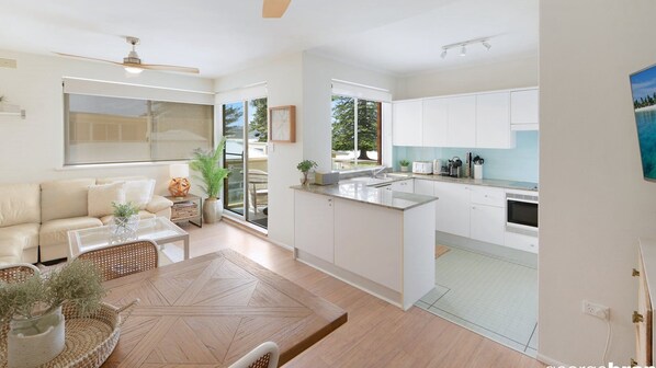 A modern kitchen with white cabinets, a dining area with a wooden table, and a living room with a beige sofa.