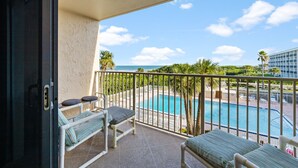 A balcony with two lounge chairs, a pool, and a view of the ocean and a building.