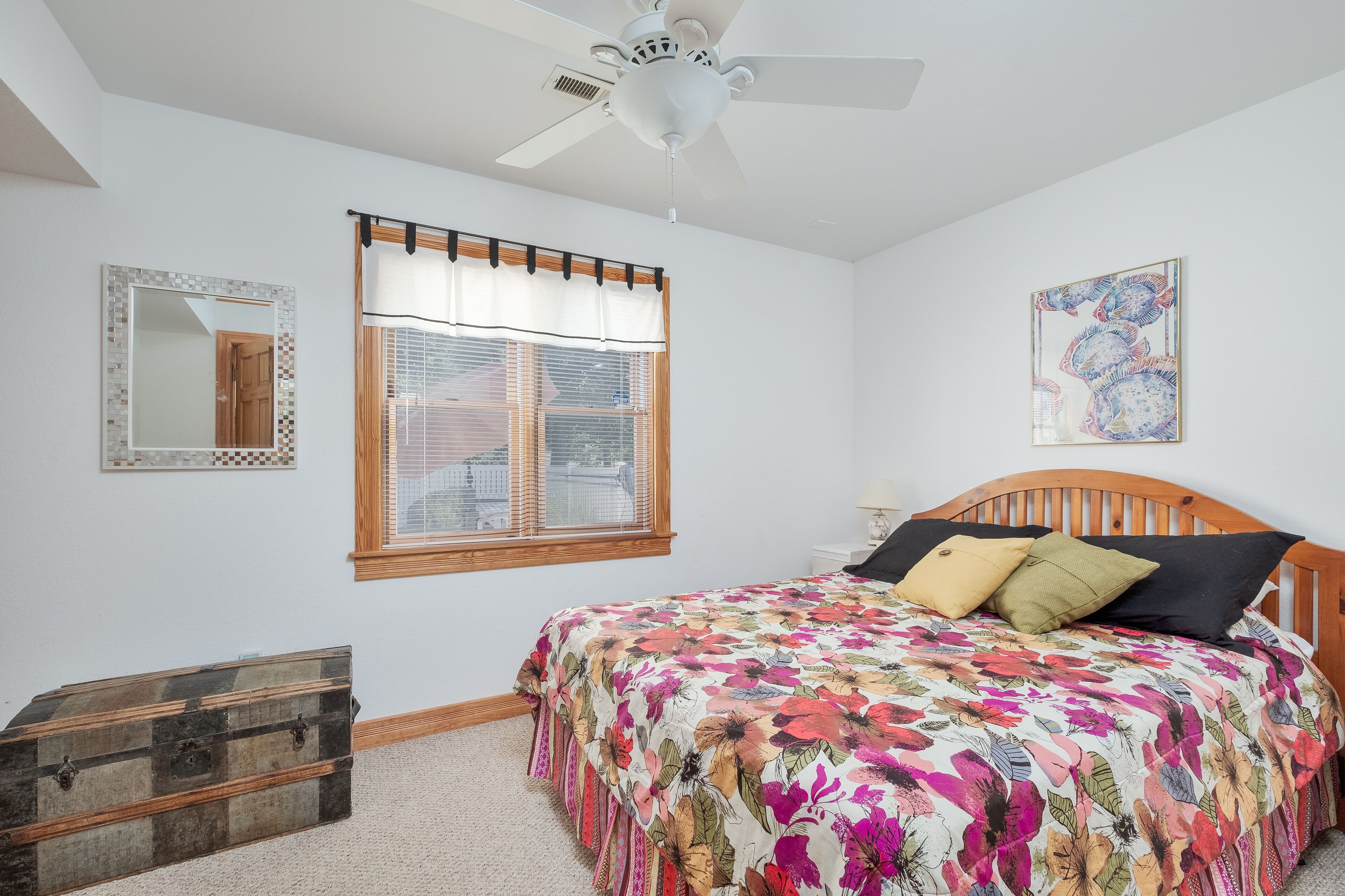 A bedroom with a wooden headboard, a floral bedspread, a ceiling fan, a window with blinds, a mirror, and a small wooden chest.