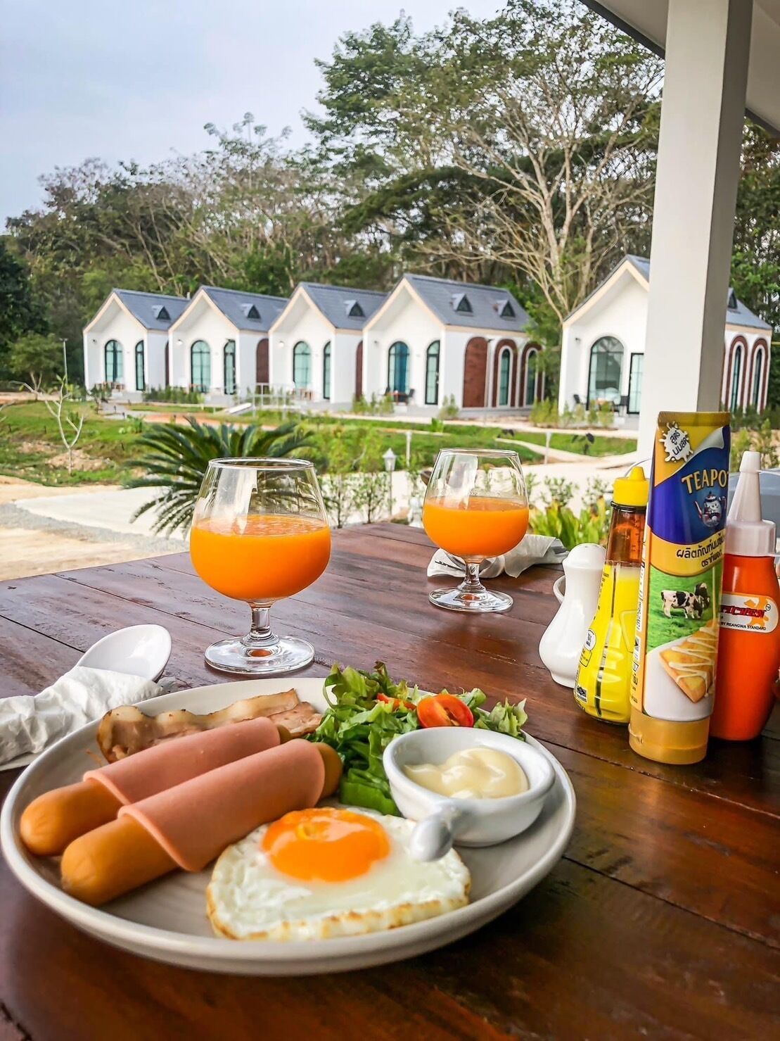 A breakfast table with eggs, sausages, and juice glasses, set against a view of a white building with arched windows.