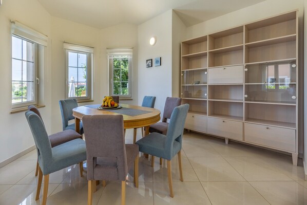 A dining area with a round table, six chairs, and a fruit bowl. There are three windows with blinds, a large bookshelf, and a tiled floor. - Villa Lotus 13F with private pool and jacuzzi. (Pula)