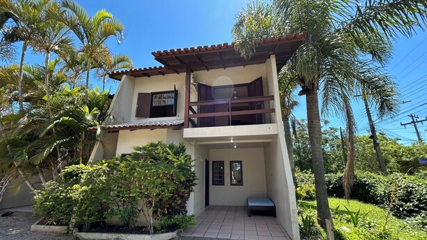 A two-story house with a balcony, surrounded by lush greenery and palm trees. - Residencial Villa Luana (Garopaba)
