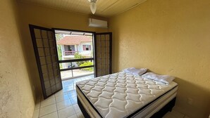 A bedroom with a bed, a window with a view of a house, and an air conditioning unit on the wall. - Residencial Villa Luana (Garopaba)