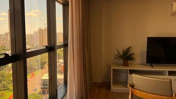 Una habitación con ventanas amplias, un televisor sobre un mueble, una silla y una planta.