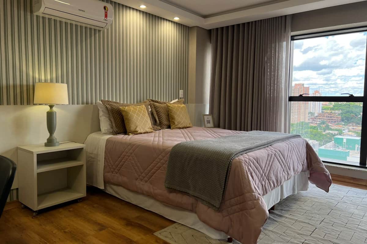 Una habitación de hotel moderna con una cama grande, una mesita de noche con lámpara, aire acondicionado integrado y vistas a la ciudad a través de un ventanal amplio.