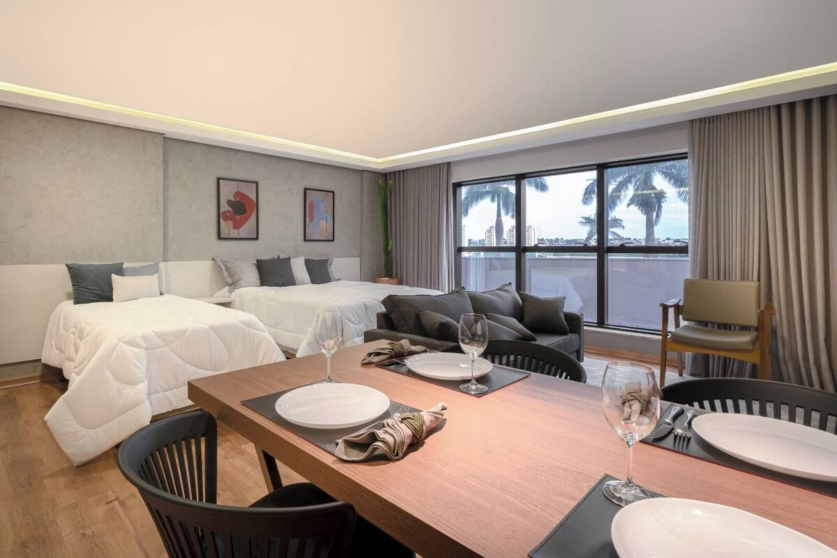 Habitación de hotel con dos camas, zona de comedor con mesa dispuesta para dos personas y vistas a palmeras y edificios a través de amplias ventanas.