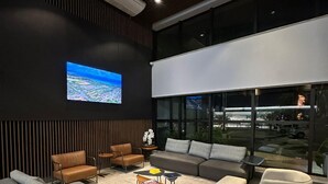 A modern lounge area with a large flat-screen TV, comfortable seating, and a view of the city through large windows. - Hotel Flat Afonso Pena by FGibran (Campo Grande)