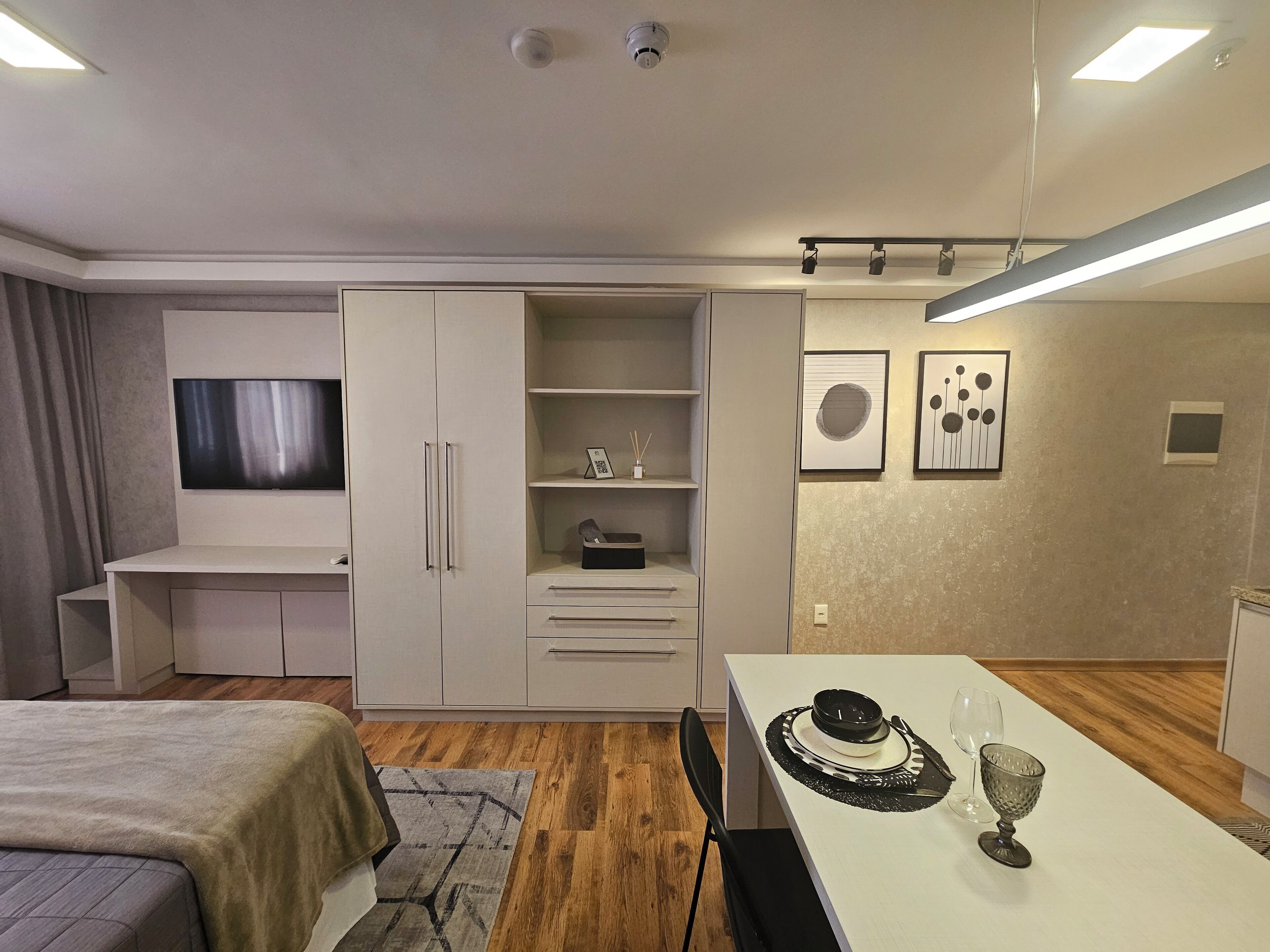 Quarto moderno com cama, televisão, guarda-roupa, secretária e área de refeições com mesa posta para dois.