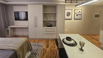 Quarto moderno com cama, televisão, guarda-roupa, secretária e área de refeições com mesa posta para dois.