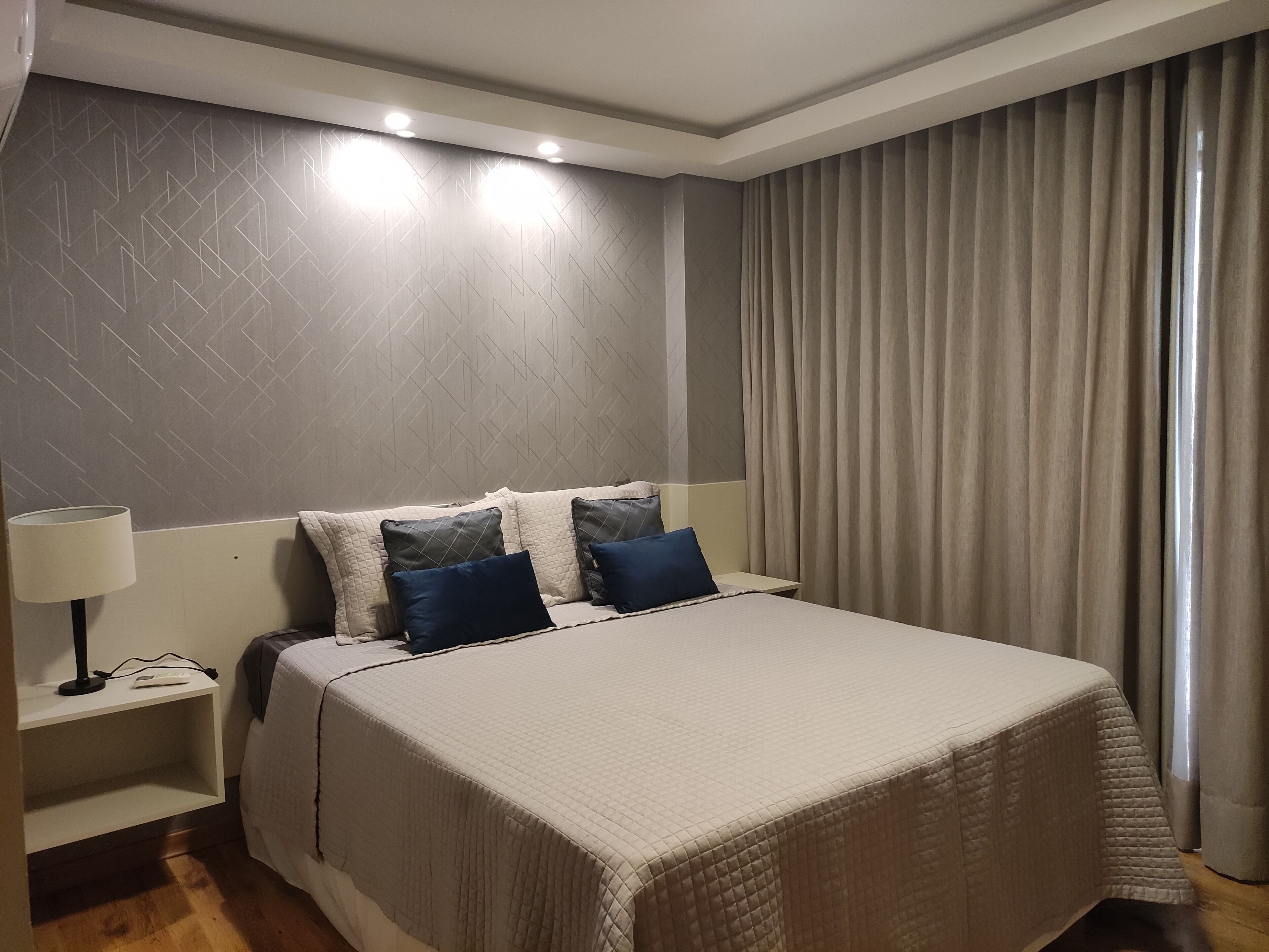 Quarto de hotel com cama, mesa de cabeceira, luminária e janela com cortinas.