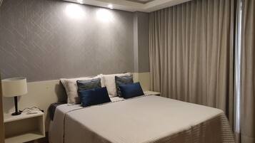 Quarto de hotel com cama, mesa de cabeceira, luminária e janela com cortinas.