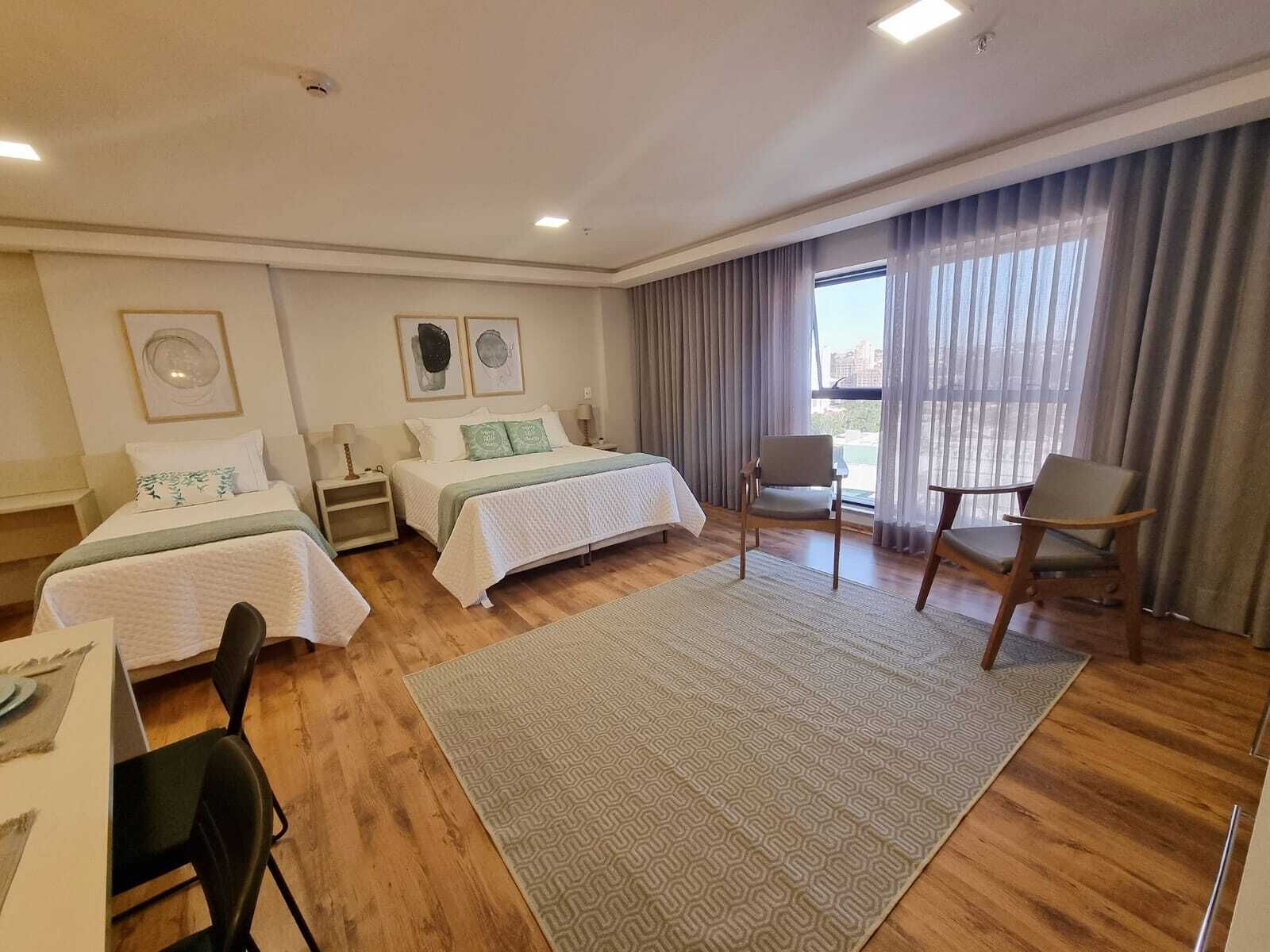 A hotel room with two beds, a dining table, chairs, a large window with curtains, and a view of the outdoors.
