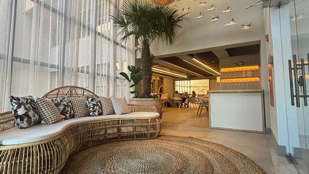 A spacious lobby with a large woven rug, a curved seating area with cushions, and a tall palm tree. - Hotel Flat Afonso Pena by FGibran (Campo Grande)