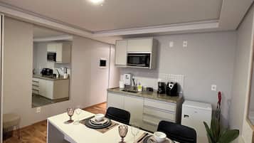 Cocina compacta con armarios blancos, microondas y refrigerador. Una mesa de comedor con sillas está colocada en el centro.