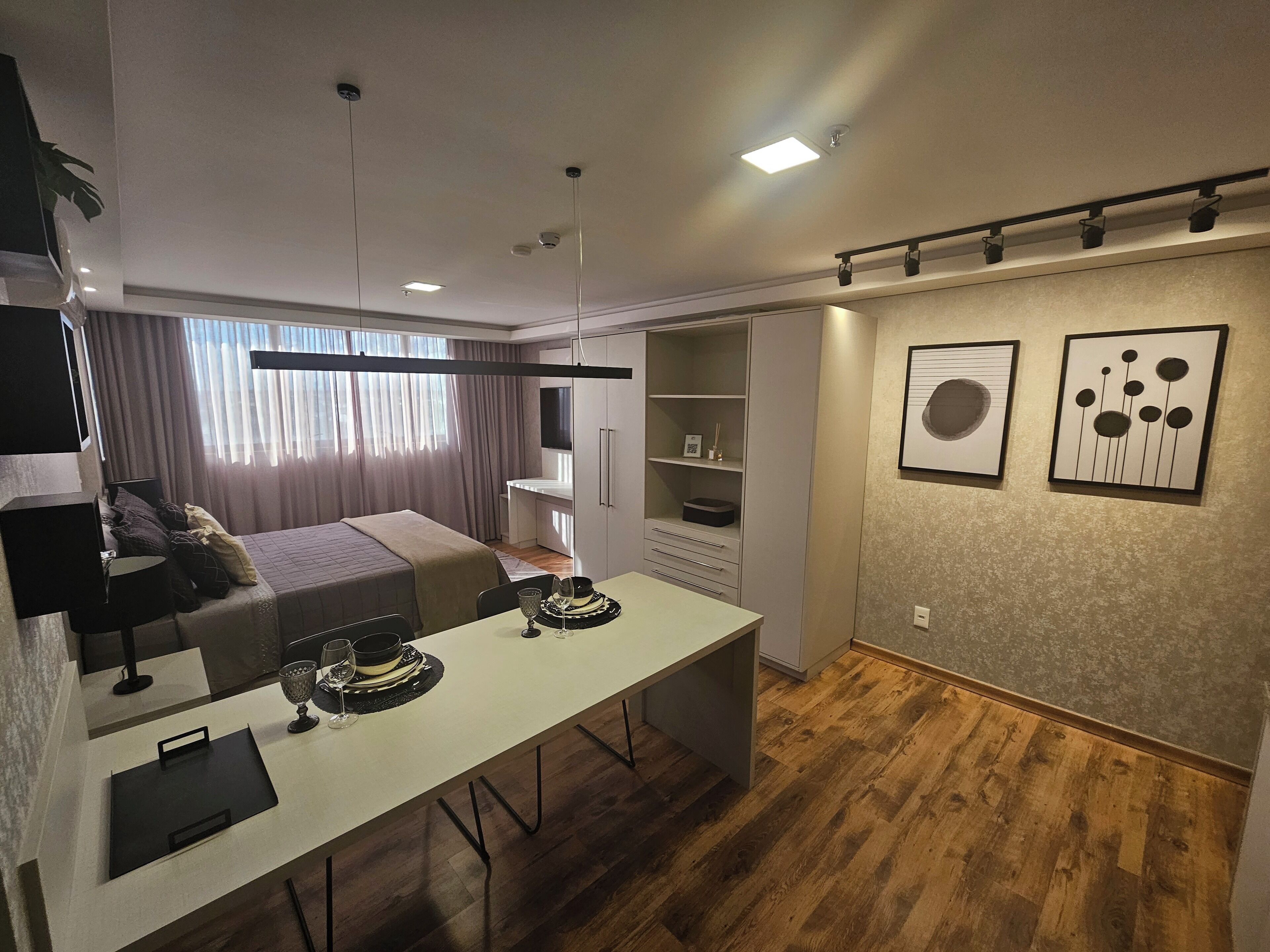 A modern hotel room with a bed, bedside table, desk, and chairs. There is a television on the wall, a bookshelf, and artwork on the wall.