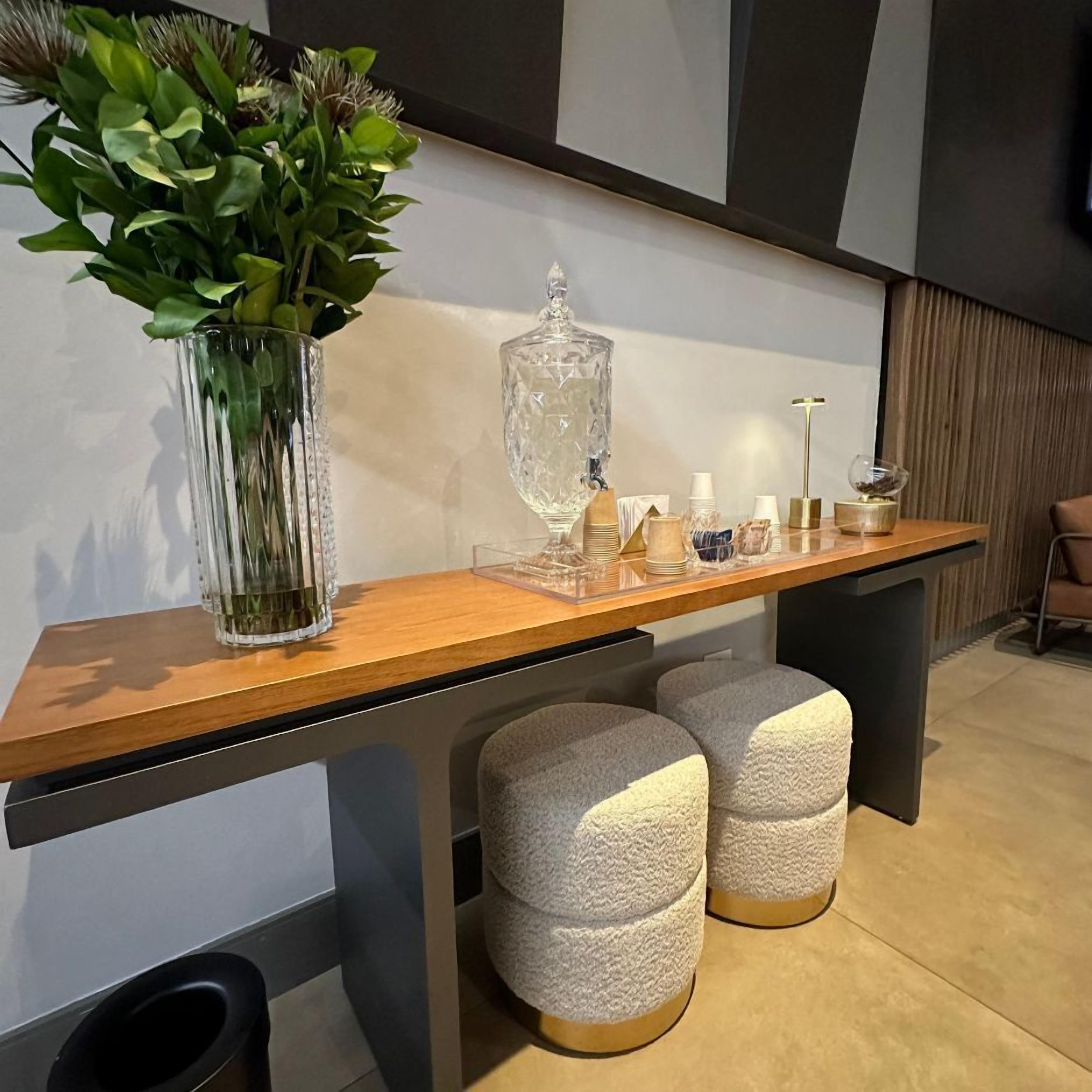 A modern bar area with a wooden countertop, a glass vase with green plants, a crystal decanter, and two textured stools.