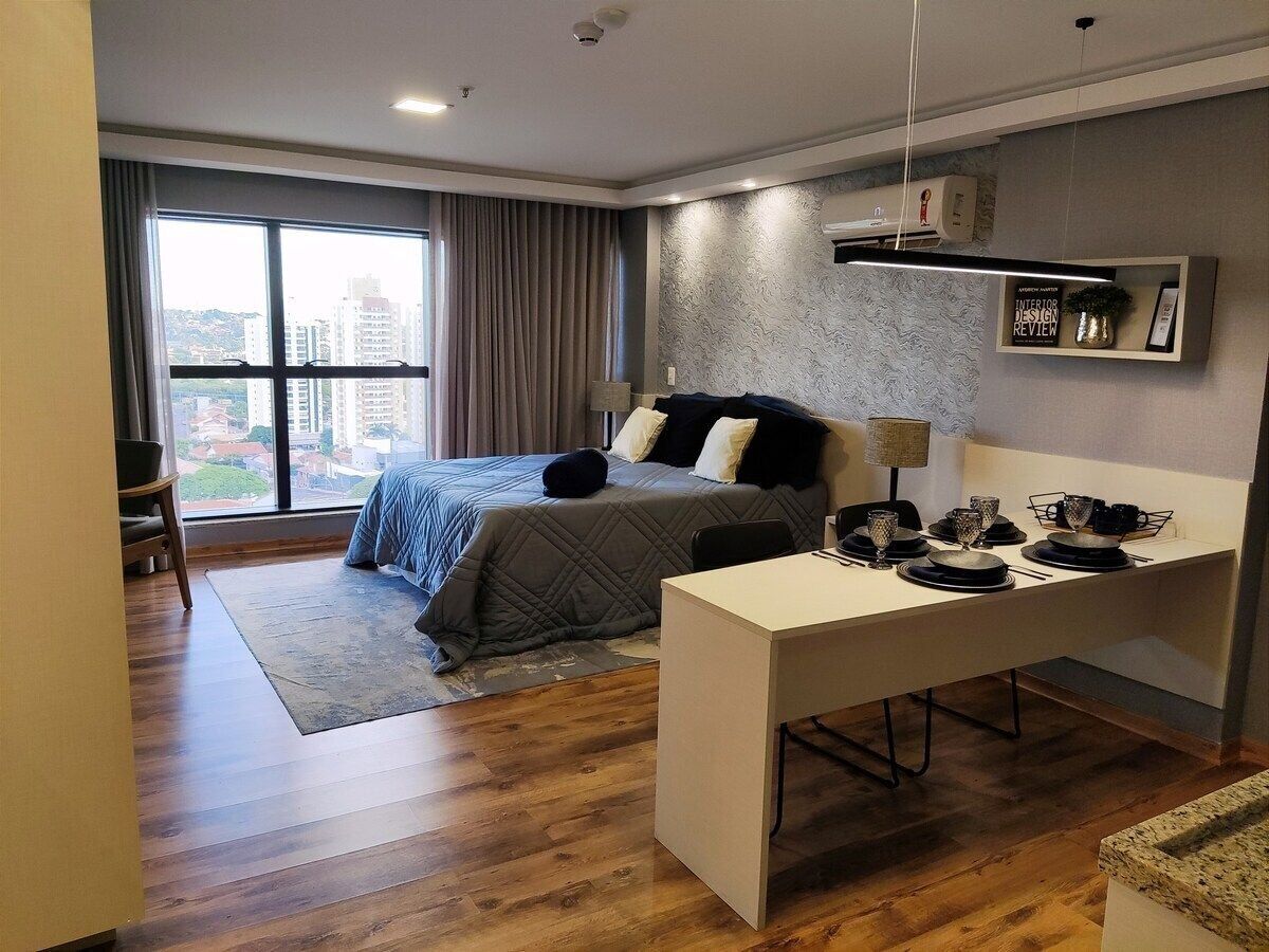 A modern hotel room with a large bed, a desk, and a view of the city through large windows.