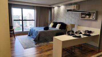 A modern hotel room with a large bed, a desk, and a view of the city through large windows.