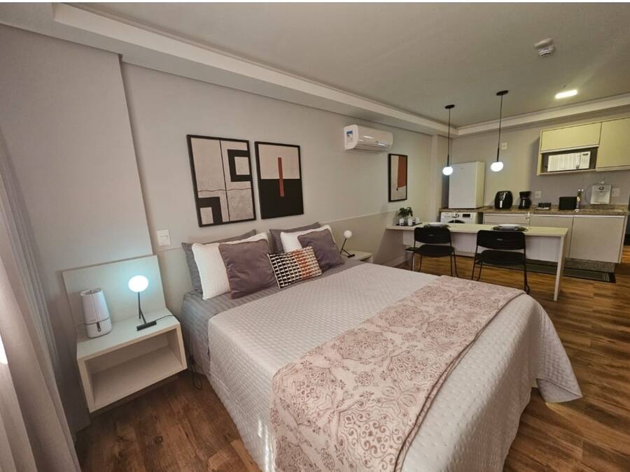 A modern bedroom with a large bed, bedside tables, a desk, and a kitchenette.