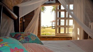 Una cama con almohadones florales de colores, una colcha blanca y vistas a un área verde con palmeras a través de una ventana con cortinas blancas.