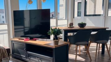 Een moderne woonkamer met een flatscreen televisie, een houten salontafel, een potplant en een eethoek met stoelen.
