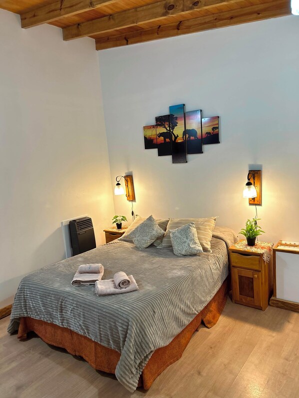 Una cama bien hecha con almohadas, una mesita de noche con lámpara, una obra de arte en la pared y un radiador.