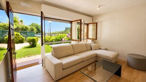 Een moderne woonkamer met een beige modulaire bank, een glazen salontafel en uitzicht op een tuin en heuvels.