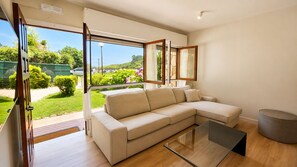 A modern living room with a beige sectional sofa, a glass coffee table, and a view of a garden and hills.