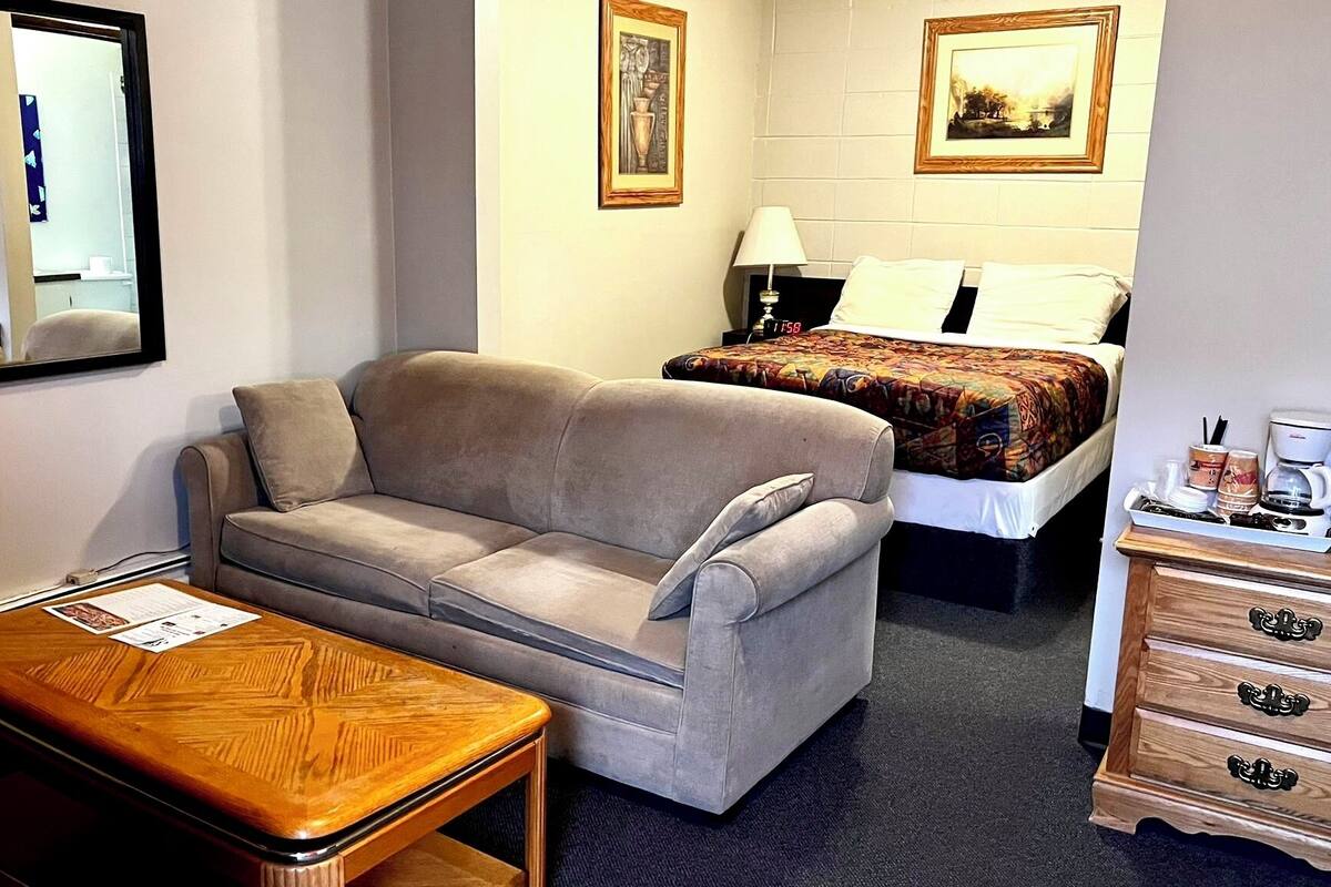 A hotel room with a sofa, a wooden side table, a bed with a colorful blanket, a dresser with drawers, and two framed pictures on the wall.