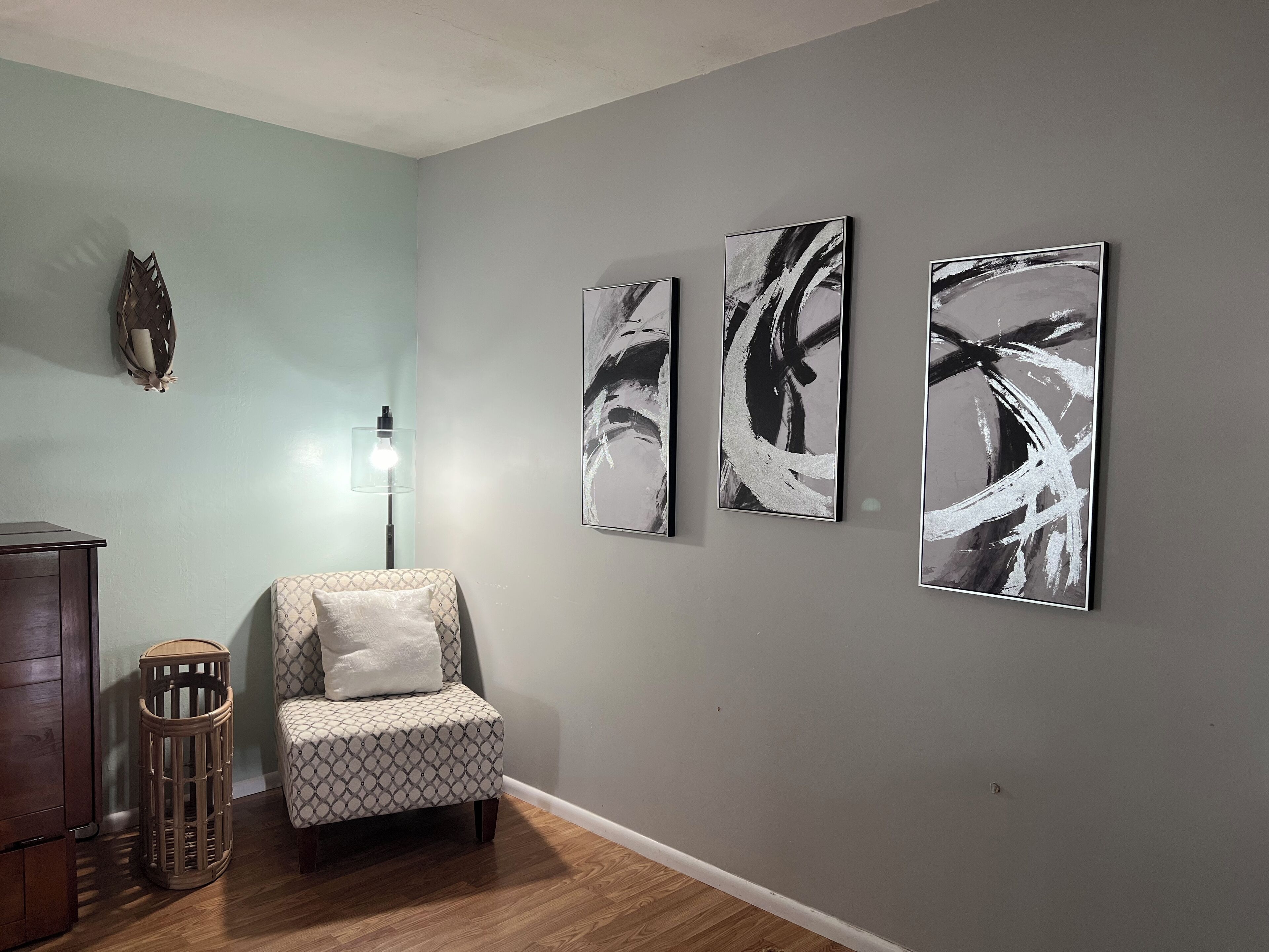 Une pièce comprenant un fauteuil à motifs, une petite table d’appoint en bois et trois œuvres d’art abstrait accrochées au mur.