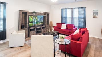 Un salon avec un téléviseur, un canapé rouge, un fauteuil blanc et un ventilateur de plafond.