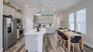 A modern kitchen with a central island, a dining area with a wooden table and chairs, and a sign above the island that reads "Each House". - Spring Break, Sunset Deck, Gameroom, 1 block to beach, Sleeps 17, gas grill (Panama City Beach)