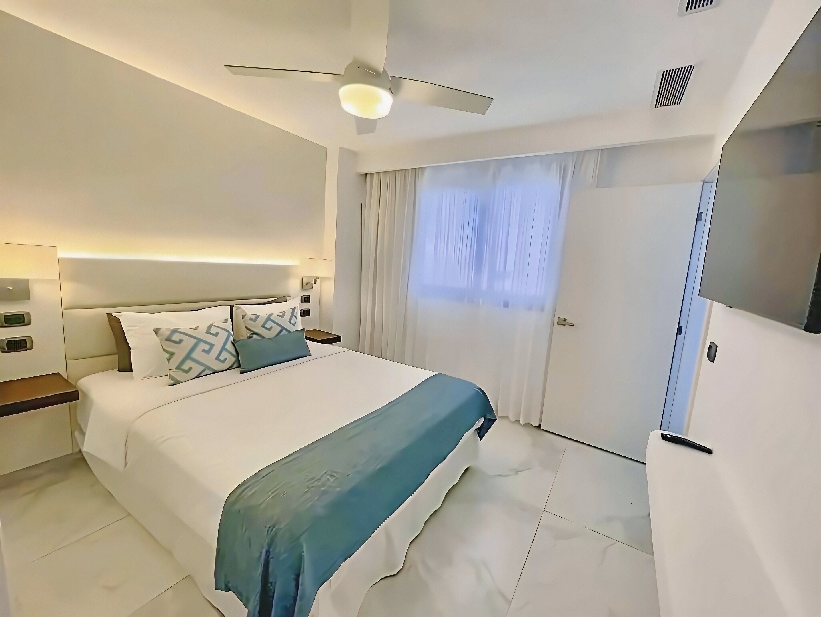 A modern hotel room with a bed, bedside tables, a ceiling fan, a TV, and a door.